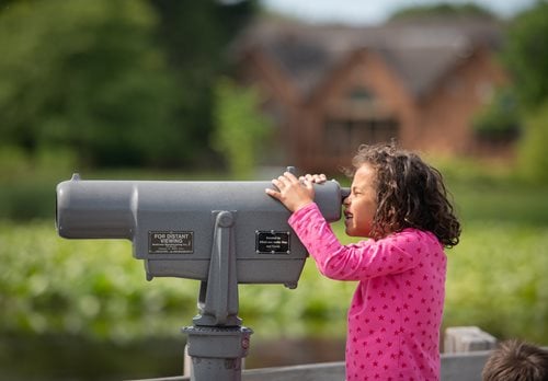  young girl peers through a telescope, focused on the tranquil lake in front of her.