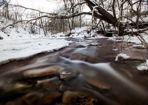A picturesque stream meanders through a snowy forest, flanked by rocks and trees, embodying a peaceful winter scene.